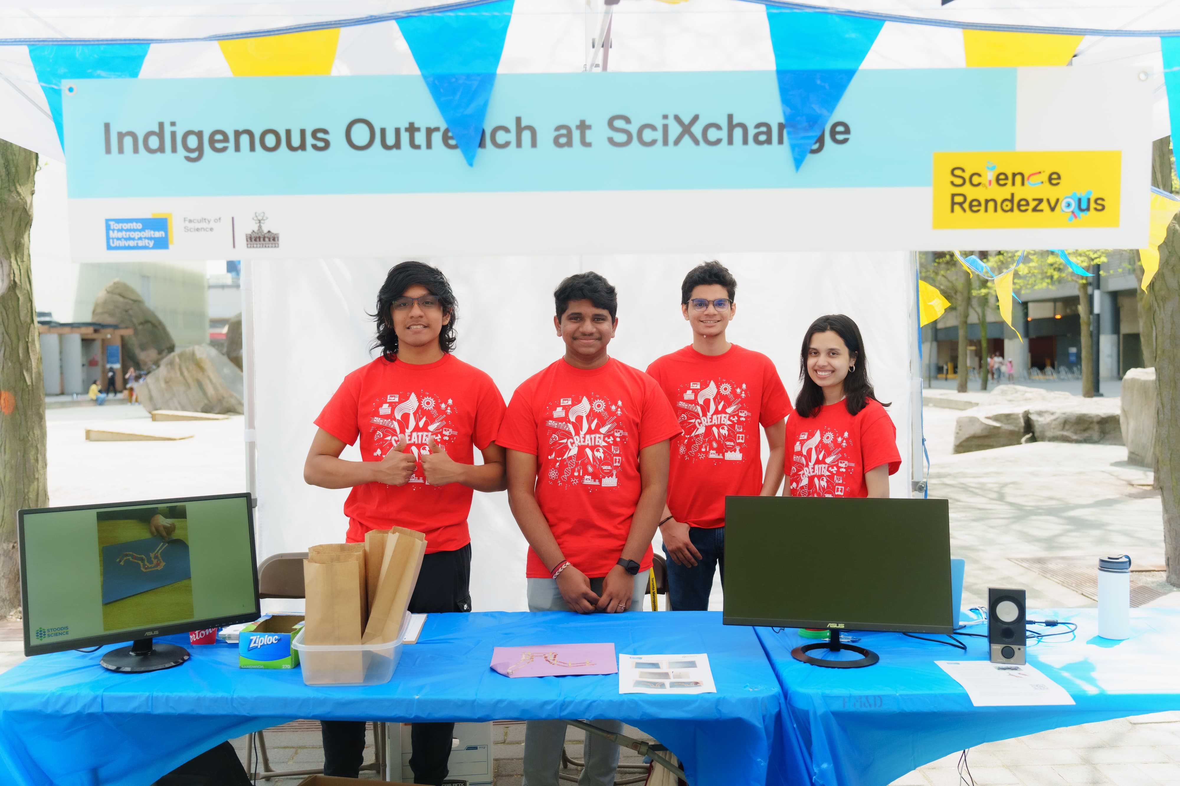 Four TMU volunteers stand at a booth under a banner saying "Indigenous Outreach at SciXchange"
